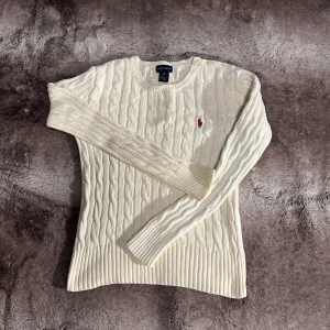 Ralph Lauren - Säljer en stilren vit stickad tröja från Ralph Lauren med kabelstickat mönster och en broderad röd logotyp på bröstet. Tröjan har långa ärmar och rund halsringning. Perfekt för kyligare dagar!
