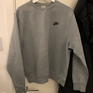 Grå sweatshirt från Nike - Säljer en stilren grå sweatshirt från Nike med deras klassiska logga på bröstet. Tröjan har långa ärmar och en rund halsringning, perfekt för en avslappnad look. Passar bra till både jeans och träningsbyxor.