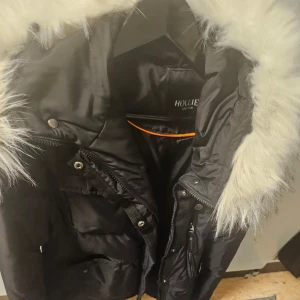Svart pufferjacka från Hollies - Snygg svart pufferjacka från Hollies med en fluffig vit fuskpäls på huvan. Jackan har dragkedja och knappar framtill för extra skydd mot kylan. Perfekt för kalla vinterdagar.