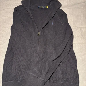 Mörkblå tröja med dragkedja från Polo Ralph Lauren - Snygg mörkgrå tröja från Polo Ralph Lauren med dragkedja och broderad logga på bröstet. Perfekt för en stilren och avslappnad look, men också för finare tillställningar. 