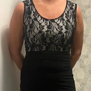 Svart klänning med spetsdetaljer - Elegant svart klänning med vackra spetsdetaljer på överdelen. Klänningen har en tight passform och är ärmlös, vilket ger en stilren look. Perfekt för speciella tillfällen.