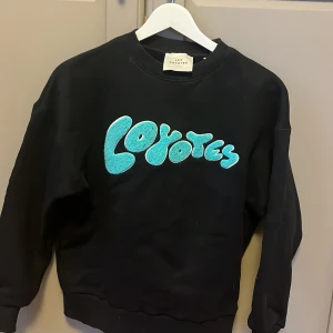 Svart sweatshirt från Les Coyotes de Paris - Snygg svart sweatshirt från Les Coyotes de Paris med turkos text 'Coyotes' på framsidan. Storlek xxs men har passat mig som är S .Perfekt för en avslappnad stil med långa ärmar och rund halsringning. Lite colleage tröja vibe, och väldigt snygg 