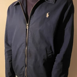 Marinblå jacka från Ralph Lauren - Säljer en stilren marinblå jacka från Ralph Lauren med dragkedja och klassisk krage. Jackan har ett broderat logotypmärke på bröstet och två framfickor. Insidan har ett rutigt mönster i blått och gult. Perfekt för en avslappnad och snygg look. Nypris 1900kr!