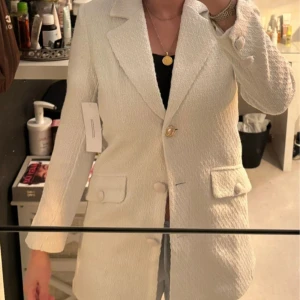 Vit kavaj  - Elegant vit kavaj med knappar framtill och klassisk krage. Kavajen har en strukturerad yta och två fickor med lock. Perfekt för en stilren look.