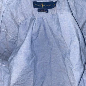 Blå skjorta från Ralph Lauren - Snygg blå skjorta från Ralph Lauren i slim fit. Skjortan har en broderad logga på bröstet och knappar framtill. Perfekt för en stilren look.