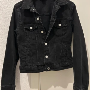 Svart jeansjacka med silvriga knappar - Snygg svart jeansjacka med silverfärgade knappar och klassisk krage. Jackan har två bröstfickor med knappstängning och en stilren design som passar perfekt till vardags. Köpt från H&M.