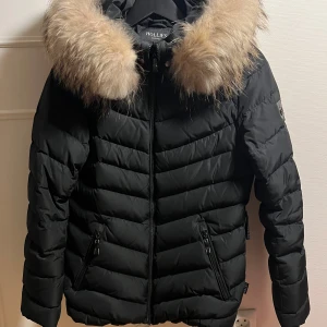 Svart dunjacka med äkta päls från Hollies - Snygg svart dunjacka från Hollies med en lyxig pälsdetalj på huvan. Jackan har en quiltad design och dragkedja framtill. Insidan är fodrad med ett leopardmönster. Perfekt för kalla dagar!