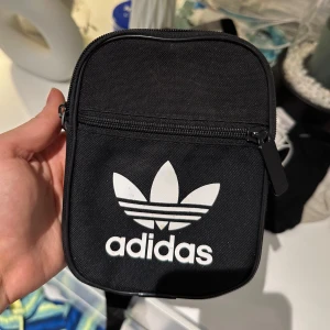 Svart axelväska från Adidas - Snygg och praktisk svart axelväska från Adidas med det klassiska treklöverlogotypen i vitt. Väskan har en dragkedja upptill och en justerbar axelrem för bekväm passform. Perfekt för att bära med sig det nödvändigaste. Helt ny!