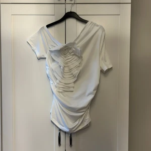 Vit asymmetrisk topp med volangdetalj - Säljer en vit asymmetrisk topp med en unik volangdetalj framtill. Toppen har en one-shoulder design och korta ärmar, vilket ger en stilren och modern look. Perfekt för att sticka ut vid speciella tillfällen.