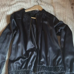 Svart jacka från Vila - Snygg svart jacka från Vila med dragkedja framtill. Jackan har en stilren design med långa ärmar och en rund halsringning. Den är i satin