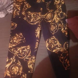 Svarta byxor med guldmönster - Snygga svarta byxor med ett iögonfallande guldmönster. Perfekta för att sticka ut med en unik stil. Byxorna har en normal passform och är tillverkade i ett bekvämt material. Från Versace jeans