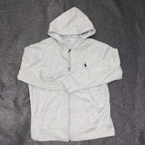Grå hoodie från Polo Ralph Lauren - Helt ny, snygg och bekväm grå hoodie från Polo Ralph Lauren med dragkedja och justerbar huva. Den har en broderad logotyp på bröstet och är perfekt för en avslappnad stil. 