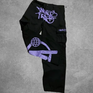 Svarta cargopants från MinusTwo - Coola svarta cargopants med lila graffiti-inspirerat tryck. Byxorna har flera fickor och en avslappnad passform. Perfekta för en streetwear-look.