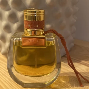 Nomade Absolu de Parfum från Chloé - Elegant parfymflaska från Chloé med en gyllene vätska. Flaskan har en unik, rundad form med en lyxig guldfärgad kork och detaljer i läderimitation. Perfekt för den som söker en sofistikerad doftupplevelse. Aldrig använts.