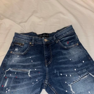 Blå jeans från Supply & Demand - Snygga blå jeans från Supply & Demand med coola färgstänk och slitna detaljer. De har en klassisk femficksdesign och knappgylf. Perfekta för en avslappnad stil med en twist.