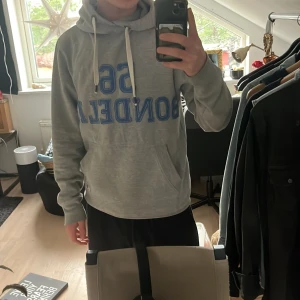 Grå hoodie från Bondelid - Säljer en grå hoodie från Bondelid med blå text '56 BONDELID' på framsidan. Tröjan har en klassisk känguruficka och justerbar huva med vita snören. Perfekt för en avslappnad stil.