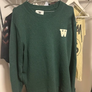 Grön stickad tröja Wood Wood - Snygg grön stickad tröja med en vit W-logga på bröstet. Tröjan har en rund halsringning och långa ärmar, perfekt för kyligare dagar. Passar bra till både jeans och kjol.