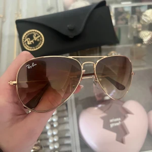 Guldiga solglasögon från Ray-Ban - Snygga guldiga Ray-Ban solglasögon i klassisk pilotmodell med bruna tonade glas. Perfekta för soliga dagar och kommer med ett stilrent svart fodral för säker förvaring. Nypris 1200kr