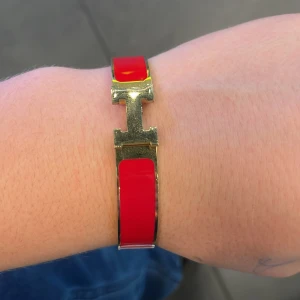 Rött armband med gulddetaljer - Snyggt och stilrent armband i röd färg med gulddetaljer. Armbandet har en elegant design med ett H-format spänne som ger det en exklusiv look. Perfekt accessoar för att lyfta vilken outfit som helst, oavsett om det är till vardags eller fest.