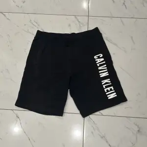 Snygga svarta shorts från Calvin Klein med deras logga tryckt i vitt längs ena benet. Perfekta för en avslappnad stil eller träning. De är i ett mjukt och bekvämt material som känns skönt mot huden. Passar perfekt för sommarens varma dagar eller en svettig träningspass. 🖤