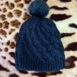 Svart stickad mössa från H&M - Säljer en superfin svart stickad mössa från H&M med en fluffig boll på toppen. Perfekt för kalla dagar och ger en mysig känsla. Den har ett snyggt flätmönster och ribbad kant. Passar perfekt till både vardag och vinteräventyr! 🖤
