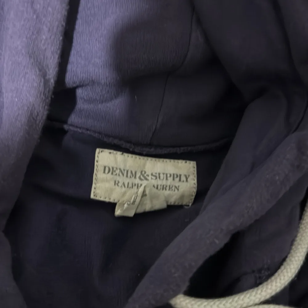 Säljer en snygg mörkblå hoodie från Ralph Lauren, perfekt för en avslappnad stil. Den har en stor ficka fram och en justerbar huva med snören. Texten 'DNM & SUPPLY' är tryckt i gult på framsidan. Passar perfekt för höst och vinter! Lila blå färg. Tröjor & Koftor.