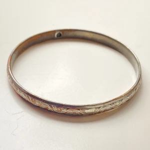 Vintage silverfärgat armband - Säljer ett vintage armband i silverfärg med ett vackert ingraverat mönster runt om. Armbandet har en klassisk och tidlös design som passar perfekt till både vardag och fest. Det är i bra skick och har en diameter på cirka 7 cm. Perfekt för dig som älskar unika smycken med en historia!