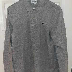 Säljer denna grå Lacoste polo-tröja, longsleeve med knappar. Då den börjar bli för liten för mig. Endast använd 2 gånger så kvalitet är mycket bra (10/10). Storlek M men är liten i storleken.