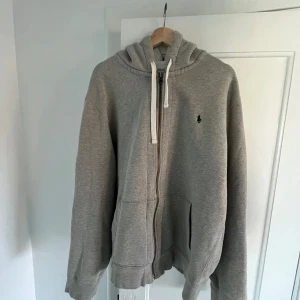 Grå hoodie från Polo Ralph Lauren - Säljer en grå hoodie från Polo Ralph Lauren i storlek XXL. Den är supermjuk och har en dragkedja framtill samt en klassisk logga på bröstet. Perfekt för kyliga dagar eller mysiga hemmakvällar. 🧥