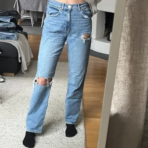 Blå jeans med slitningar - Säljer ett par snygga blå jeans med slitningar på knäna. De är högmidjade och har en straight passform som ger en avslappnad look. Köpta på gina och är utan skavanker 