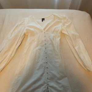 Vit långärmad klänning med knappar - Säljer en superfin vit klänning i storlek S. Den har långa ärmar och en v-ringad design med knappar framtill. Perfekt för vår och sommar! Materialet känns lätt och bekvämt, perfekt för en dag ute i solen.