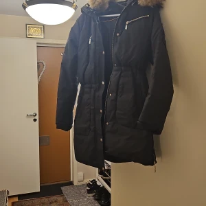 Svart parkajacka med pälsdetaljer - Säljer en svart parkajacka från Hollies i mycket bra skick. Jackan har en mysig pälsdetalj på huvan och praktiska dragkedjor. Perfekt för kalla vinterdagar. Den är långärmad och har en stilren design som passar till det mesta. Perfekt för både vardag och fest!