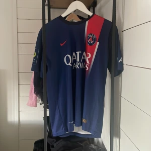 Blå PSG fotbollströja Neymar Jr - Säljer en blå PSG fotbollströja med Neymar Jr och nummer 10 på ryggen. Tröjan har korta ärmar och är tillverkad i Nike Dri-FIT material för optimal komfort. Den har en röd och vit diagonal detalj på framsidan och sponsornamnet Qatar Airways tryckt på bröstet.