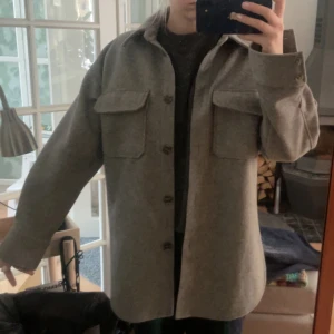 Grå overshirt med knappar - Säljer en stilren grå overshirt med knappar framtill och två stora bröstfickor. Perfekt för lager-på-lager-stil och passar bra till både vardag och mer avslappnade tillfällen.