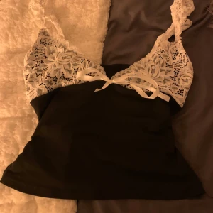 Svart linne med spetsdetaljer - Snyggt svart linne med vita spetsdetaljer och axelband. Perfekt för en stilren look med en touch av elegans. Axelbanden är justerbara och ger en feminin känsla.