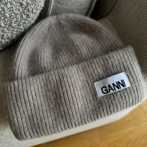 Beige stickad mössa från Ganni - Snygg beige stickad mössa från Ganni med ribbad design. Perfekt för kyliga dagar och ger en stilren look. Märkeslappen framtill ger en extra touch av stil.