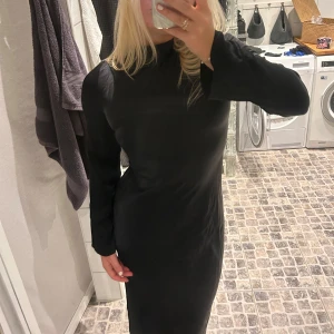 Svart långklänning med öppen rygg - Elegant svart långklänning med långa ärmar och en stilfull öppen rygg med snörningsdetaljer. Perfekt för en kväll ute eller en speciell tillställning. Klänningen har en smickrande passform och är tillverkad i ett mjukt material som ger en fin siluett.