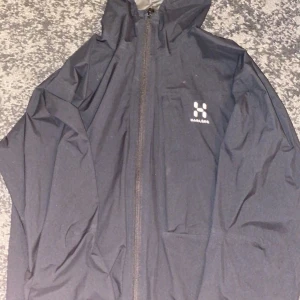 Svart vindjacka från Haglöfs - Säljer en stilren svart vindjacka från Haglöfs med dragkedja framtill. Jackan har en hög krage och är perfekt för blåsiga dagar. Den är långärmad och har en minimalistisk design med Haglöfs logga på bröstet.