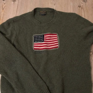 Woolrich tröja - Passar mer som M/L Säljer en olivgrön stickad tröja från Woolrich med en broderad amerikansk flagga på framsidan. Tröjan har långa ärmar och en klassisk rund halsringning. Perfekt för kyligare dagar.