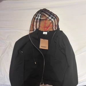 Svart hoodie från Burberry - Säljer en stilren svart hoodie från Burberry med den klassiska rutiga mönstrade huvan. Hoodien har dragkedja och en diskret broderad logga på bröstet. Perfekt för en casual look med en touch av lyx. Den är helt ny och använd 