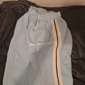 Ljusblå mjukisbyxor från Palm Angels - Säljer ett par ljusblå mjukisbyxor från Palm Angels med vita och svarta ränder längs sidorna. Byxorna har en elastisk midja med snörning och är tillverkade i polyester. Perfekta för en avslappnad stil.