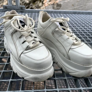 Vita sneakers från Buffalo London - Säljer ett par vita sneakers från Buffalo London med chunky sula och snörning. Skorna har en retro design med märkets logga på sidan och insidan. Perfekta för att ge lite extra höjd och stil till din outfit.