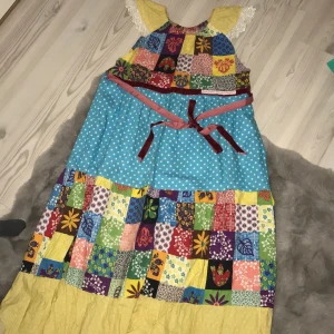 Färgglad patchwork-klänning - Unik och färgglad patchwork-klänning med ett lapptäcksmönster i olika färger och mönster. Klänningen har en mix av blommiga och prickiga mönster samt spetsdetaljer vid ärmarna. Den har en knytning i midjan och en härlig volangkant längst ner. Gjord i  Åre. Hemsydd, så finns ju bara denna klänning.