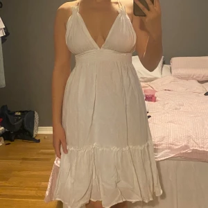 Vit sommarklänning med volangdetaljer - Säljer en söt vit klänning med v-ringad design. Klänningen är ärmlös och har en öppen rygg med knytband. Köpt på en kiosk i Dubai