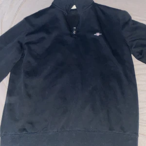 Svart tröja med dragkedja från GANT          öppen för byten !!!! - Snygg svart tröja från GANT med halv dragkedja och broderad logga på bröstet. Perfekt för en stilren look. Tröjan har långa ärmar och en hög krage.