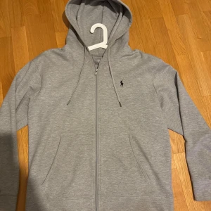 Grå hoodie från Ralph Lauren - Säljer en fin grå hoodie från Ralph Lauren med dragkedja och klassisk logga på bröstet. Tröjan har långa ärmar och en praktisk huva med dragsko. Perfekt för en avslappnad look! Hoodien ser helt ny ut och skulle säga 10/10 skick.