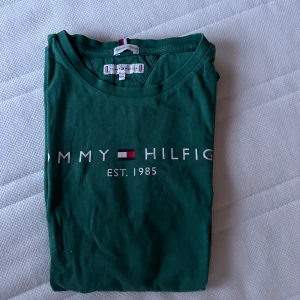 Grön t-shirt från Tommy Hilfiger - Snygg grön t-shirt från Tommy Hilfiger med tryck 'Est. 1985' på bröstet. Perfekt för en avslappnad stil med korta ärmar och rund hals. Klassisk design med märkets ikoniska logga.