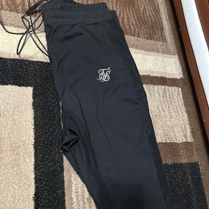 Svarta mjukisbyxor från SikSilk - Snygga svarta mjukisbyxor från SikSilk med elastisk midja och snörning. Byxorna har en stilren design med logotyp på benet. Perfekta för en avslappnad stil.