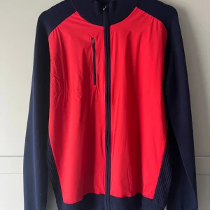 Röd och marinblå kofta med windbreaker från FootJoy  - Snygg röd och marinblå jacka från FootJoy med dragkedja framtill och en praktisk bröstficka. Perfekt för kyligare dagar med sin mix av ull och syntetmaterial. Den har långa ärmar och en stilren design med logotyp på ryggen.