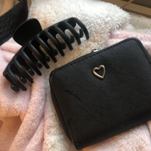 Svart plånbok med hjärtdetalj - Snygg svart plånbok med en söt hjärtdetalj på framsidan. Plånboken har dragkedja och flera fack för kort och mynt. Perfekt för att hålla ordning på dina tillhörigheter.
